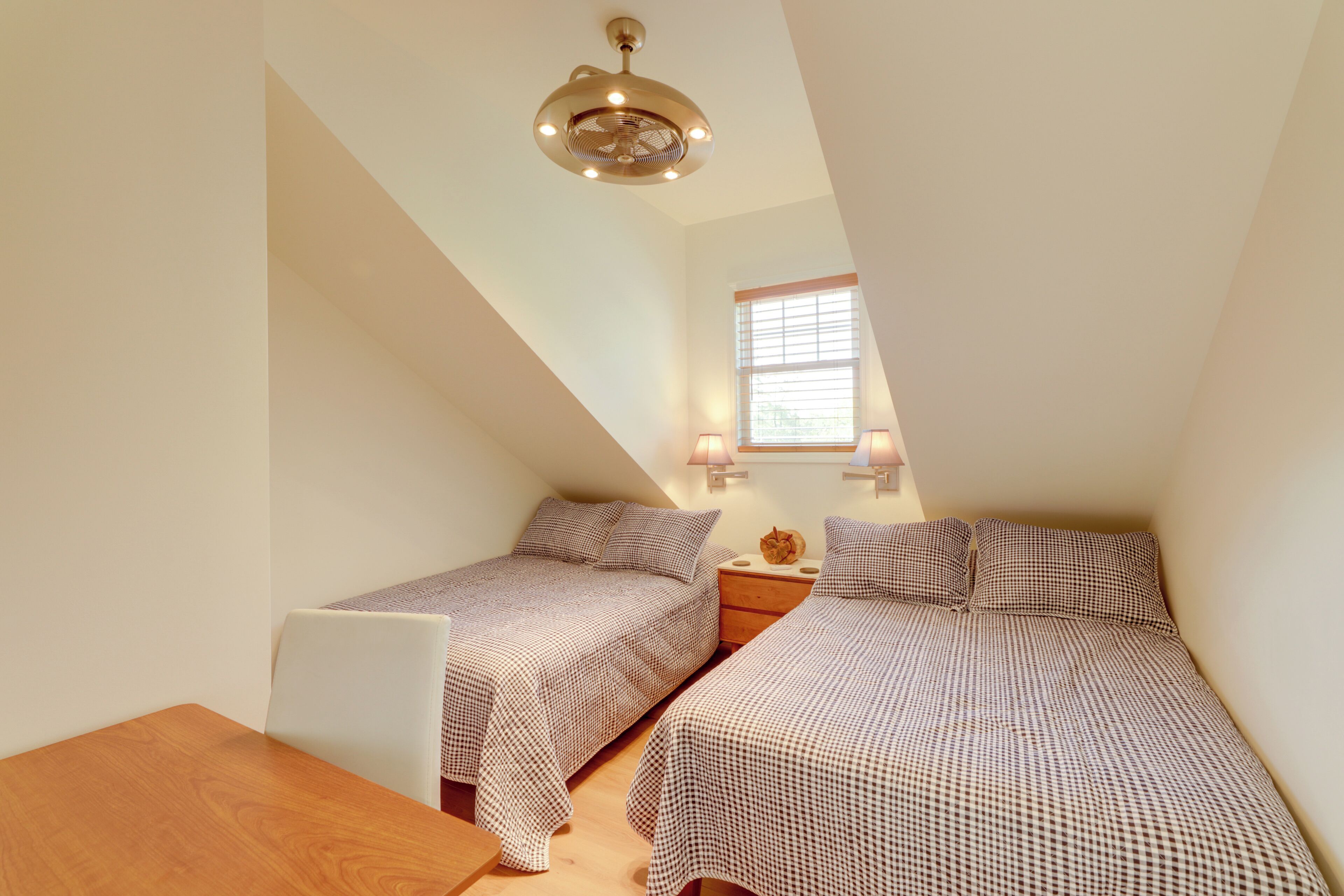 A bedroom with two beds, a wooden table, a ceiling fan, and a window with blinds.