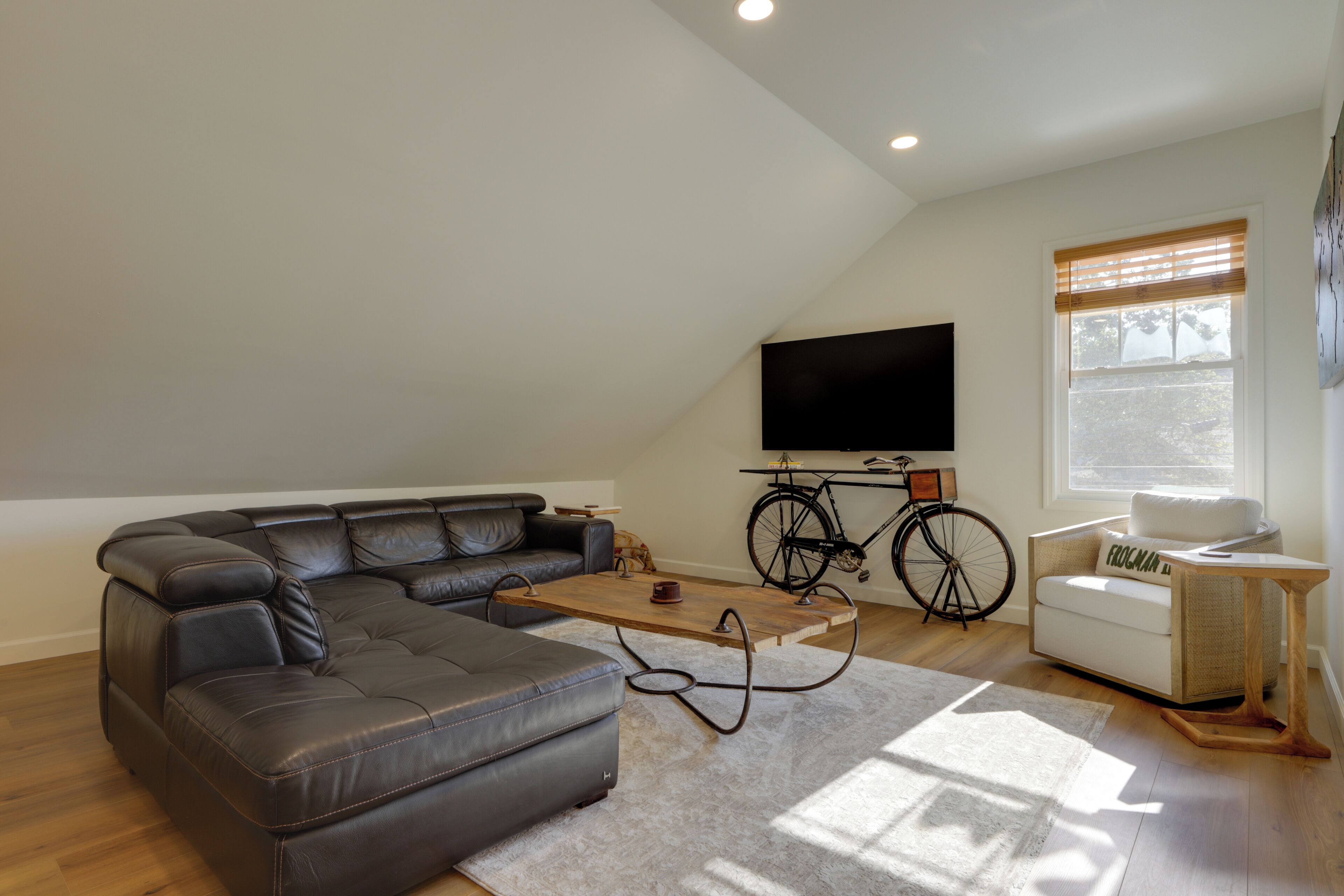 A modern living room with a sectional sofa, a coffee table, a TV mounted on the wall, a bicycle, and a window with blinds.