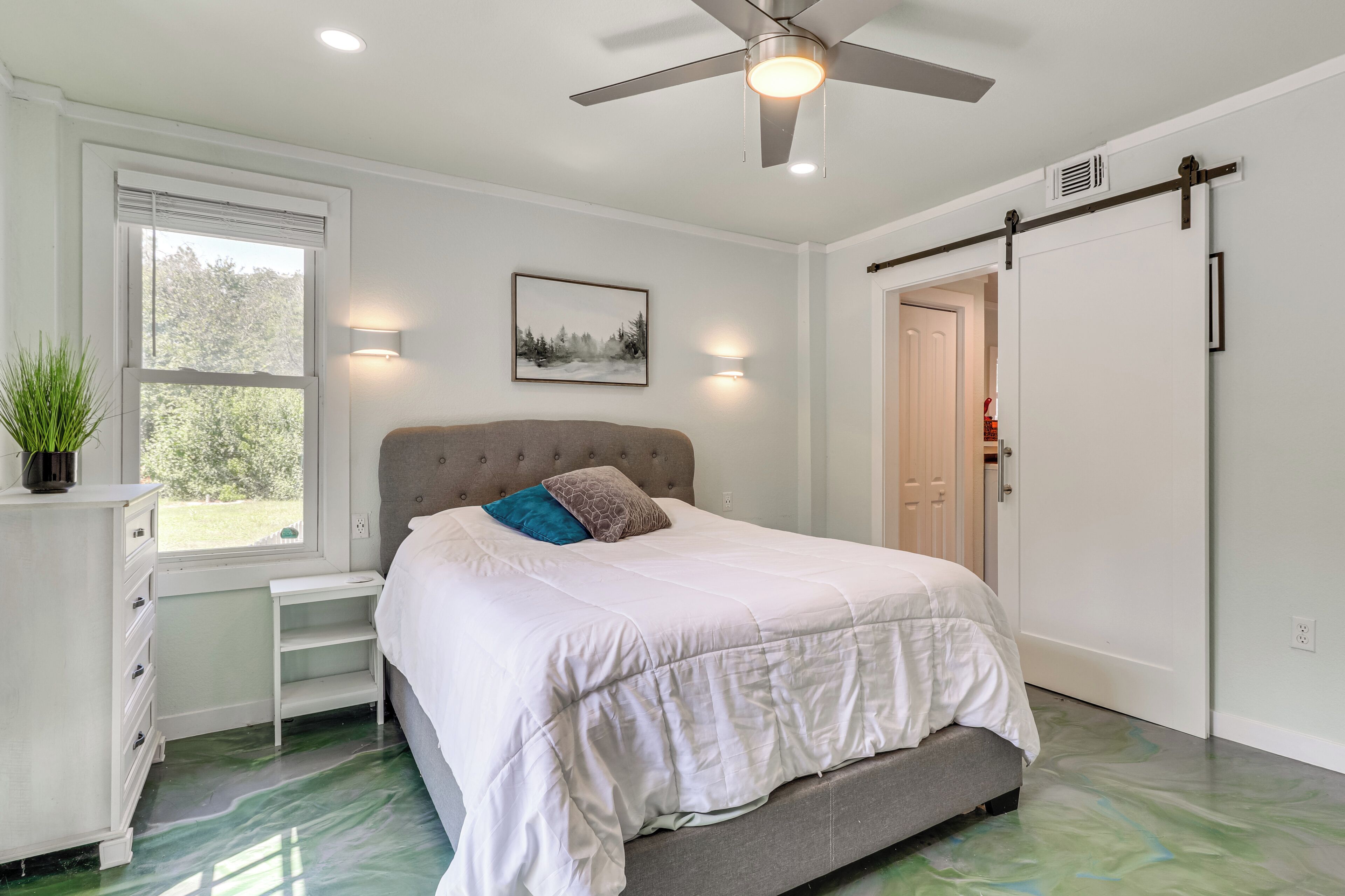 A bedroom with a bed, a ceiling fan, a window, a door, and a wall-mounted light.