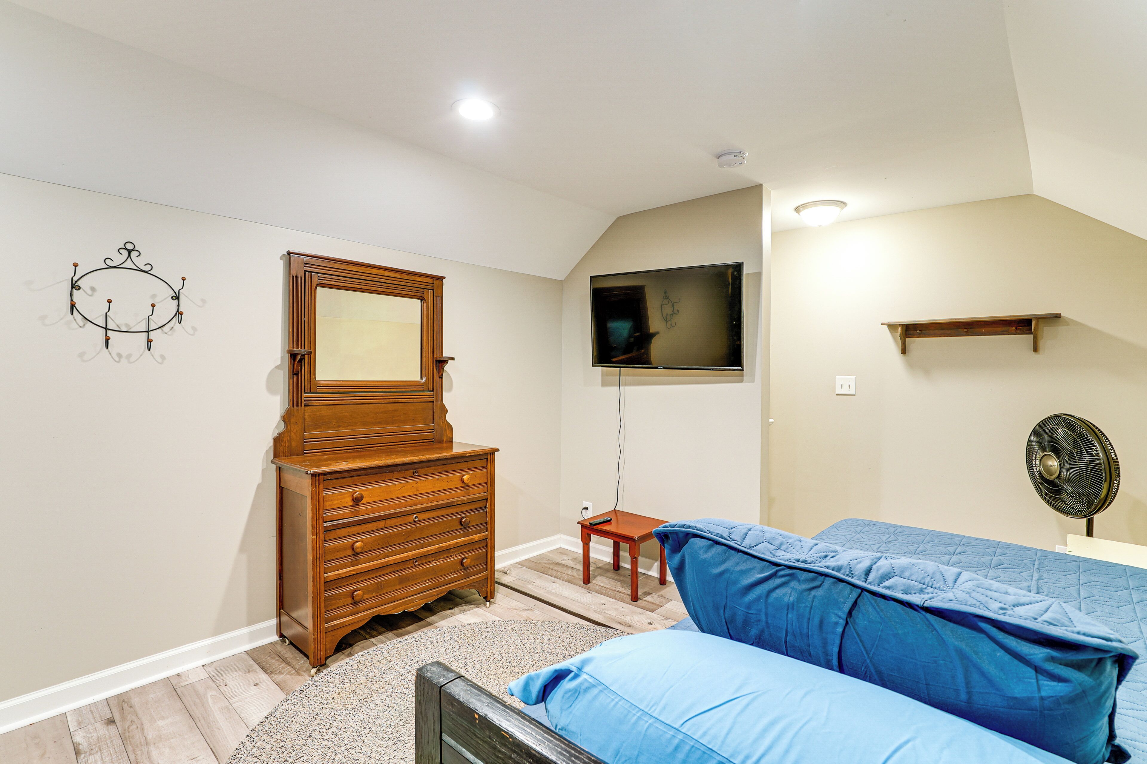 Un dormitorio con un aparador de madera, una cama con ropa de cama azul, un televisor montado en la pared y un ventilador de techo.