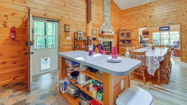 Ein Holzblockhaus-Innenraum mit Kamin aus Stein, einem Essbereich mit Tisch und Stühlen sowie einer Küchenzeile mit Regalen und Geräten.