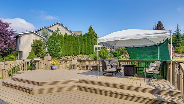 Eine Holzterrasse mit Gartenmöbeln, einem weißen Baldachin und Blick auf ein Haus und Bäume.
