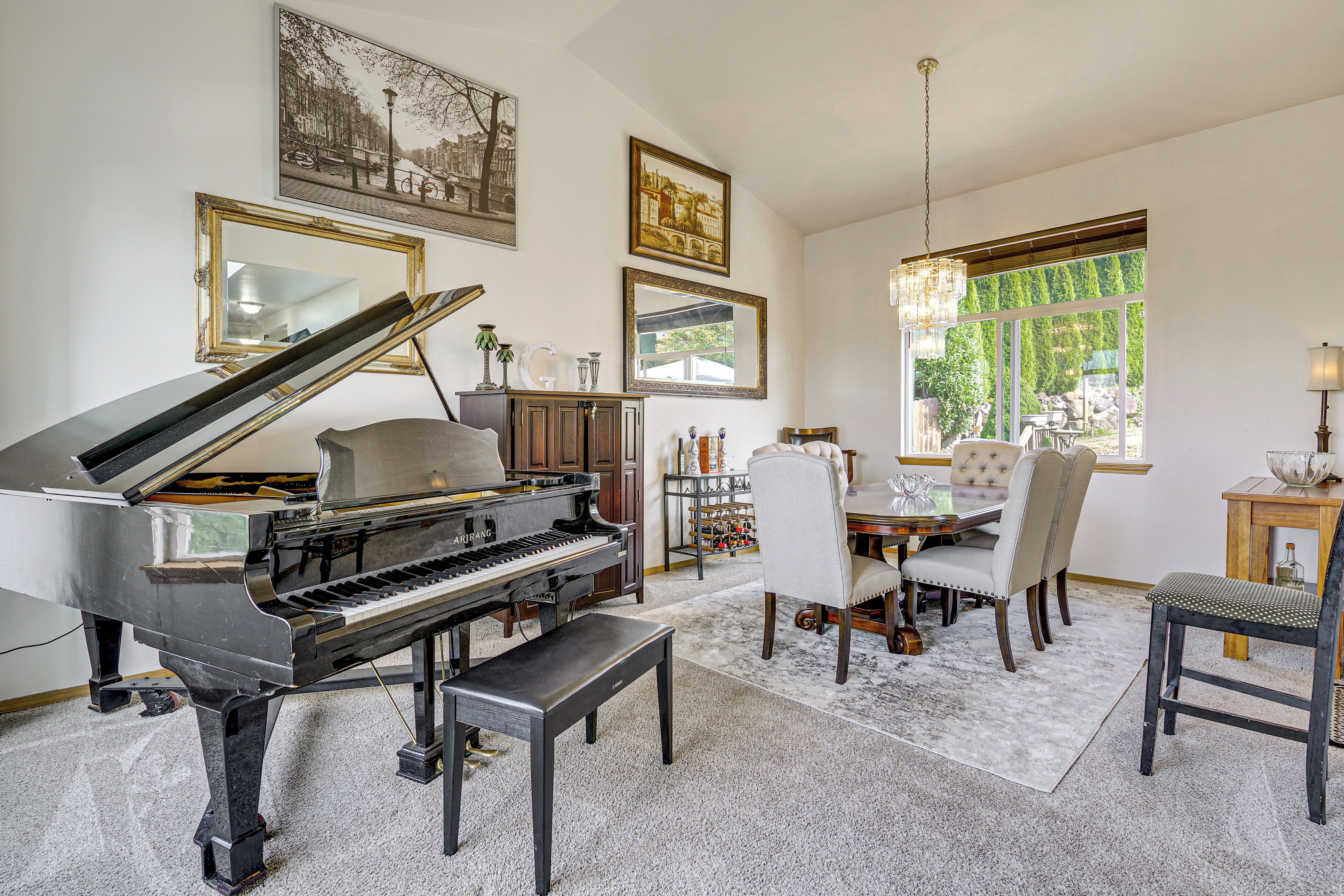 A grand piano in a spacious living room with a dining area, large windows, and artwork on the walls.