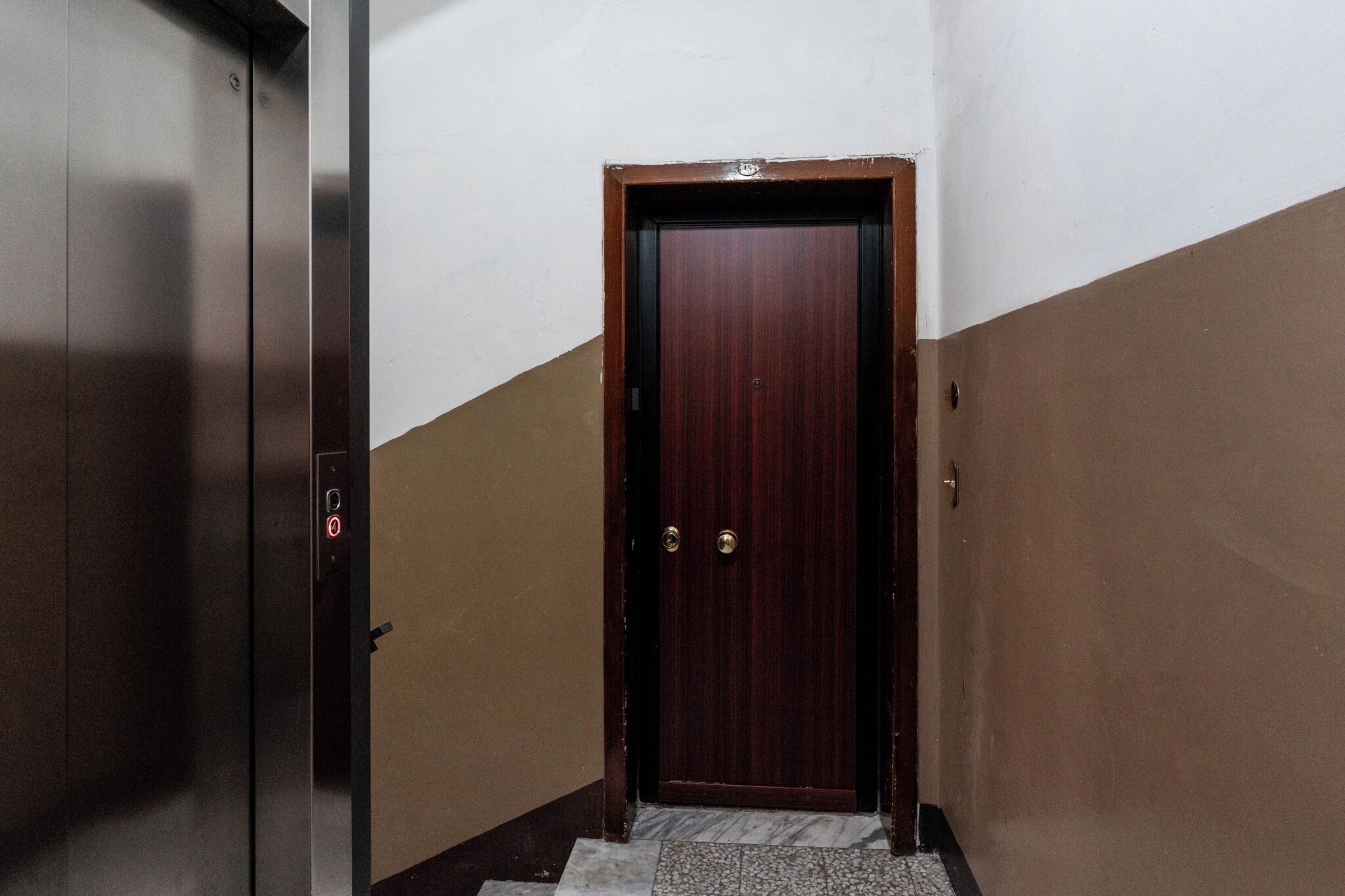 A hallway with a wooden door, a wall-mounted elevator, and a marble-tiled floor.