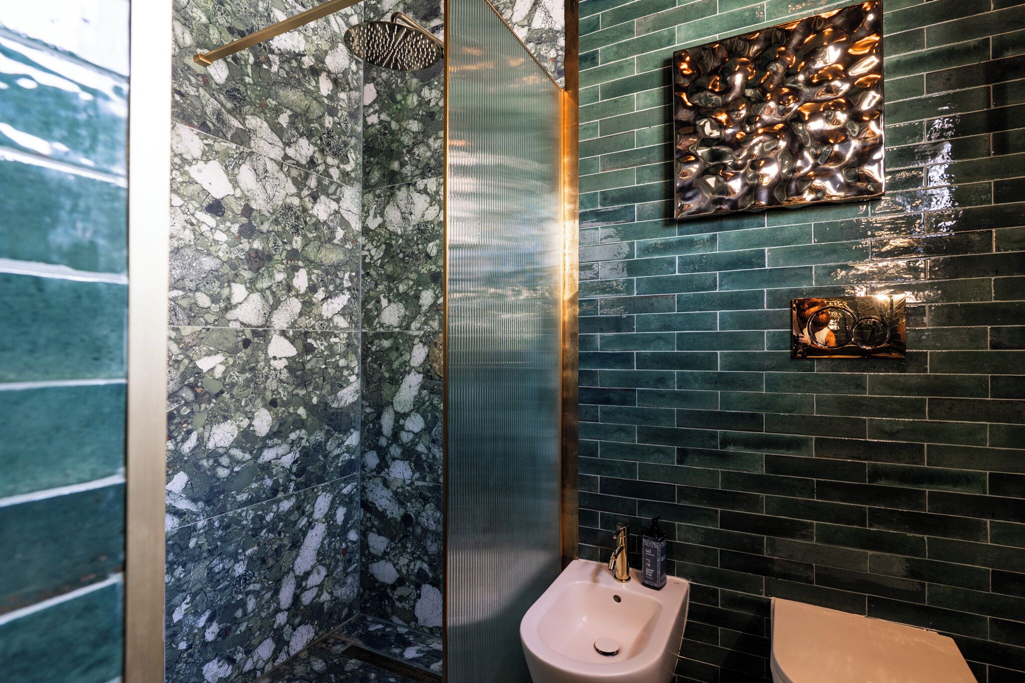 A bathroom with a marble shower, a sink with a gold faucet, and a green tiled wall with a decorative metal piece.
