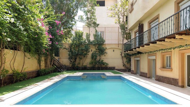 A swimming pool with a tiled edge, surrounded by a well-maintained lawn and a building with balconies. - Kennah Chill & Swim | 2BR with Pool, October City (6th of October City)