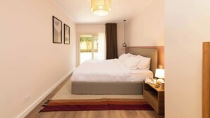 A hotel room with a bed, bedside table, lamp, and a view of the outdoors. - Kennah Chill & Swim | 2BR with Pool, October City (6th of October City)