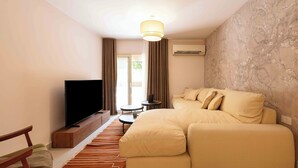 A modern hotel room with a large beige sofa, a flat-screen TV, a small round table, and a wooden bench. - Kennah Chill & Swim | 2BR with Pool, October City (6th of October City)