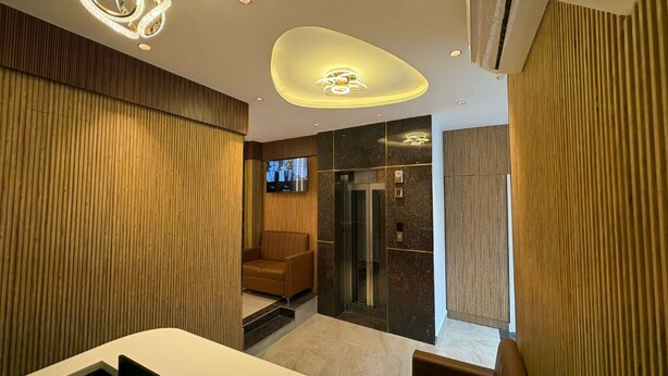 A modern hotel lobby with a wooden panel wall, a seating area, a reception desk, and a glass elevator.