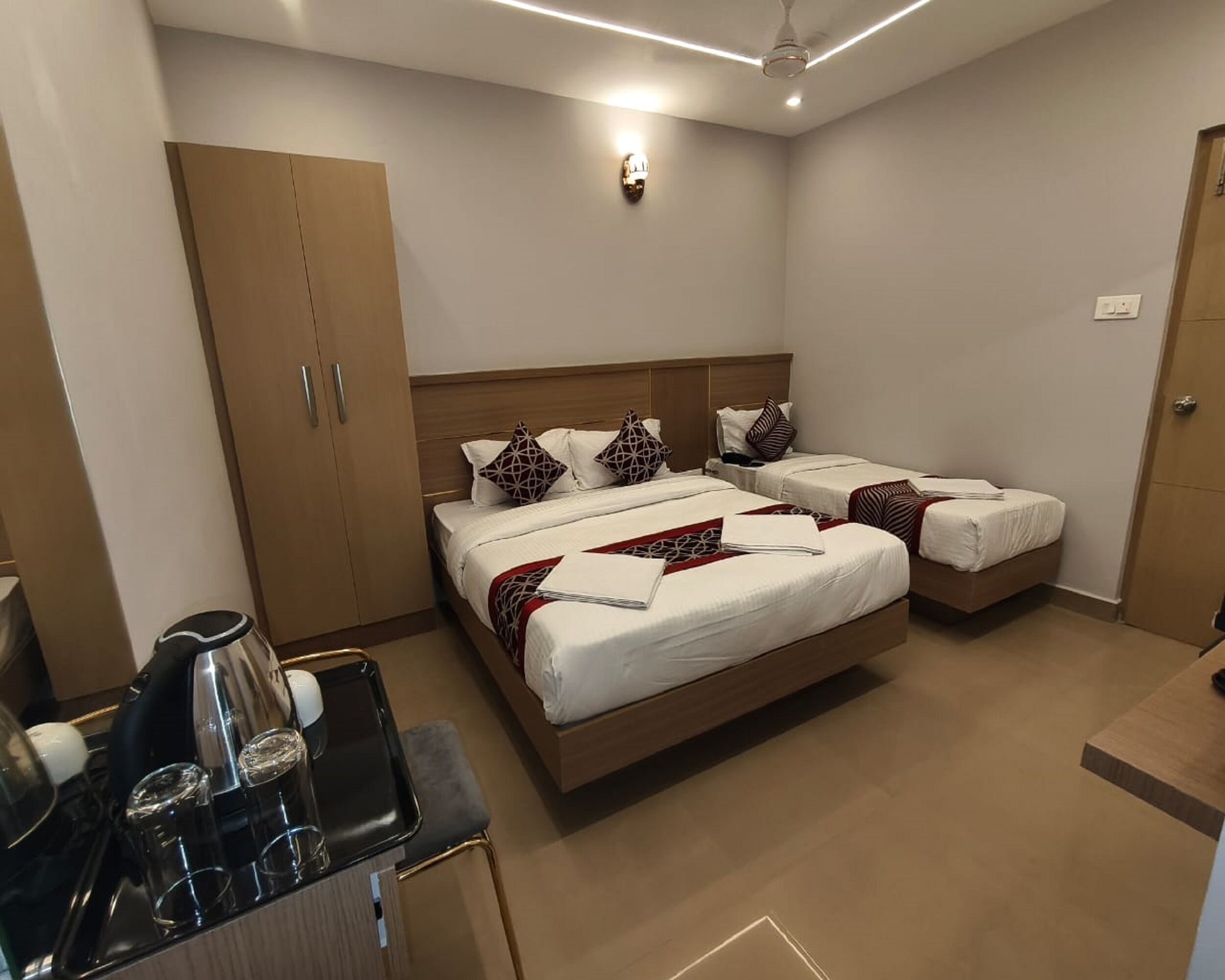 Una habitación de hotel con dos camas individuales, un armario de madera, una mesita con una tetera y vasos, y una lámpara en el techo.