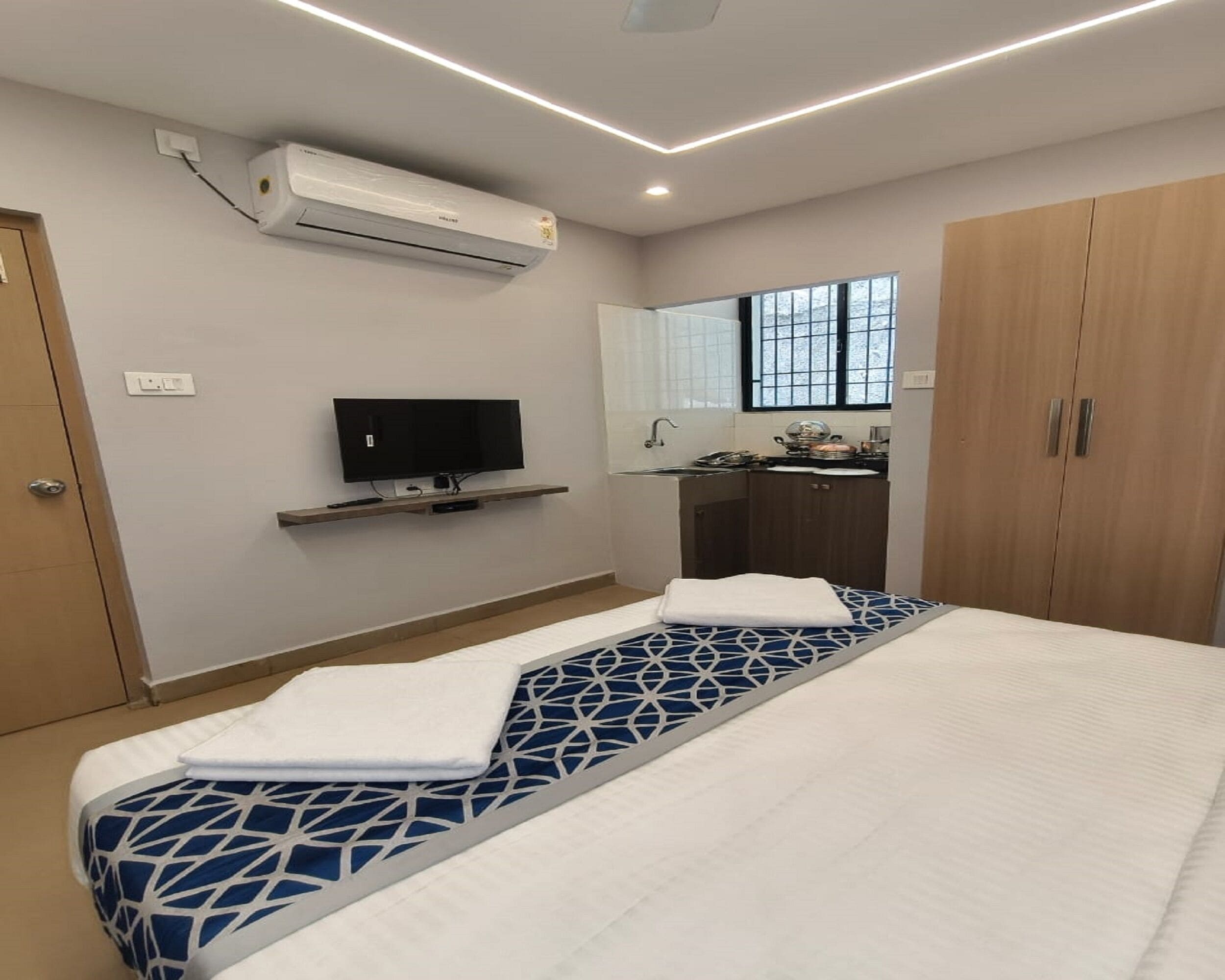 A hotel room with a bed, a TV, an air conditioner, a small kitchenette, and a wardrobe.