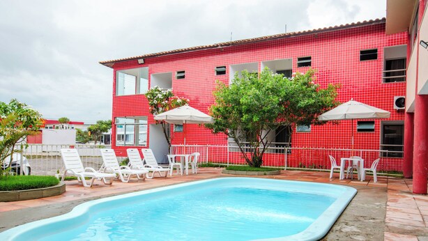 A swimming pool with white lounge chairs and umbrellas, surrounded by a red tiled building with multiple windows. - Rede Andrade Porto Praia (Porto Seguro)
