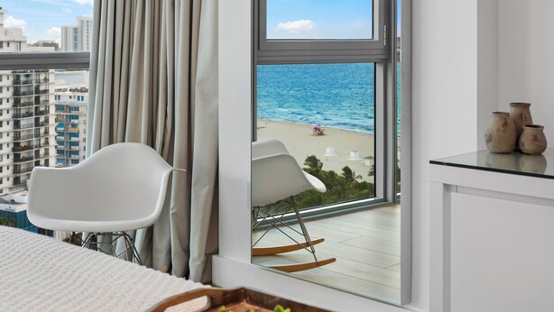 Een hotelkamer met uitzicht op het strand, een witte stoel, een schommelstoel en een kleine tafel met vazen.