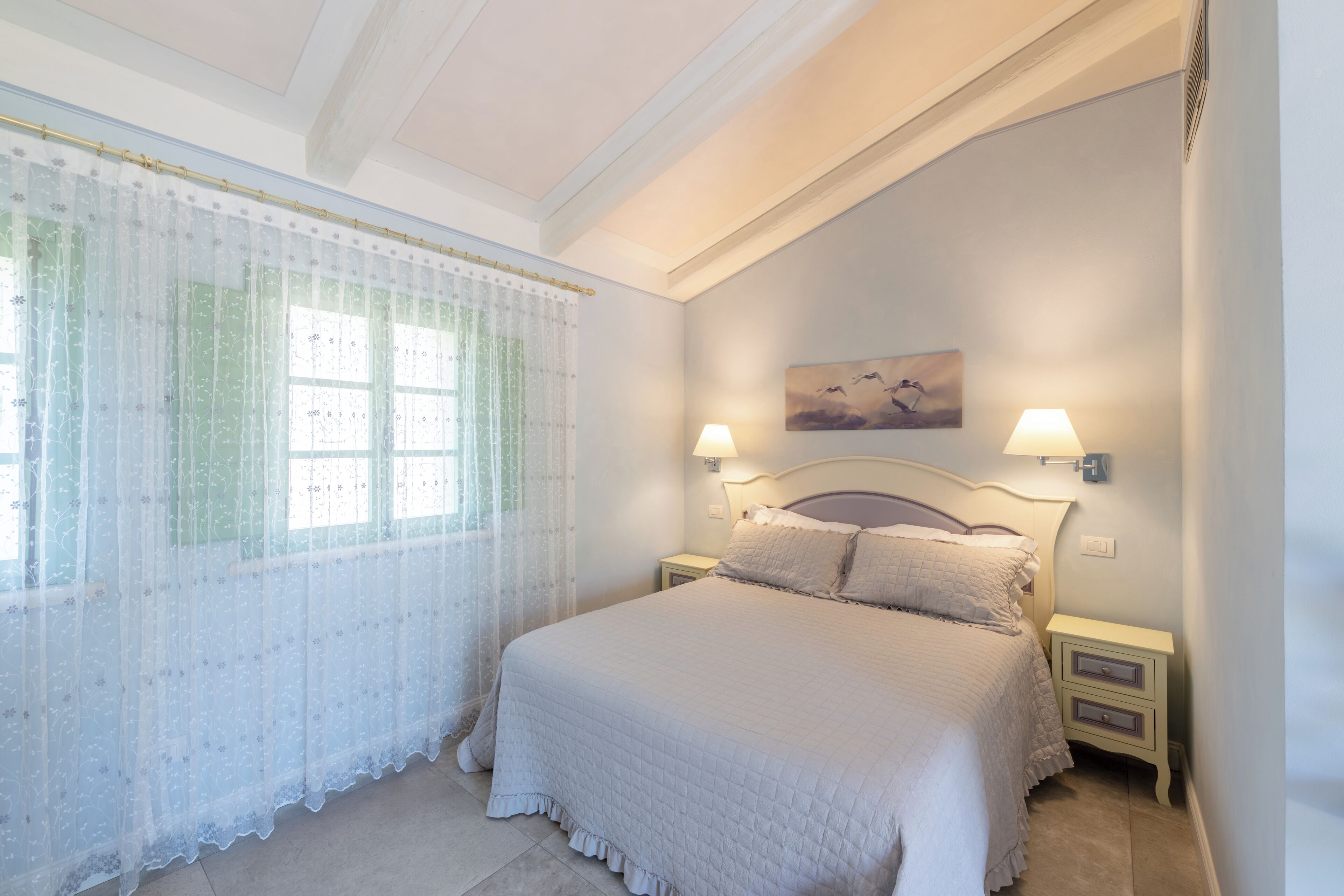 A bedroom with a large bed, two bedside tables, a window with sheer curtains, and a wall-mounted lamp.