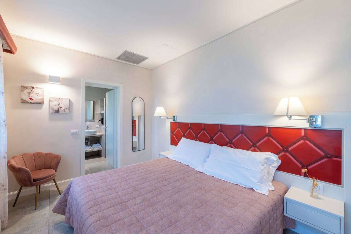 A hotel room with a bed, a chair, a vanity with a mirror, and a red headboard.