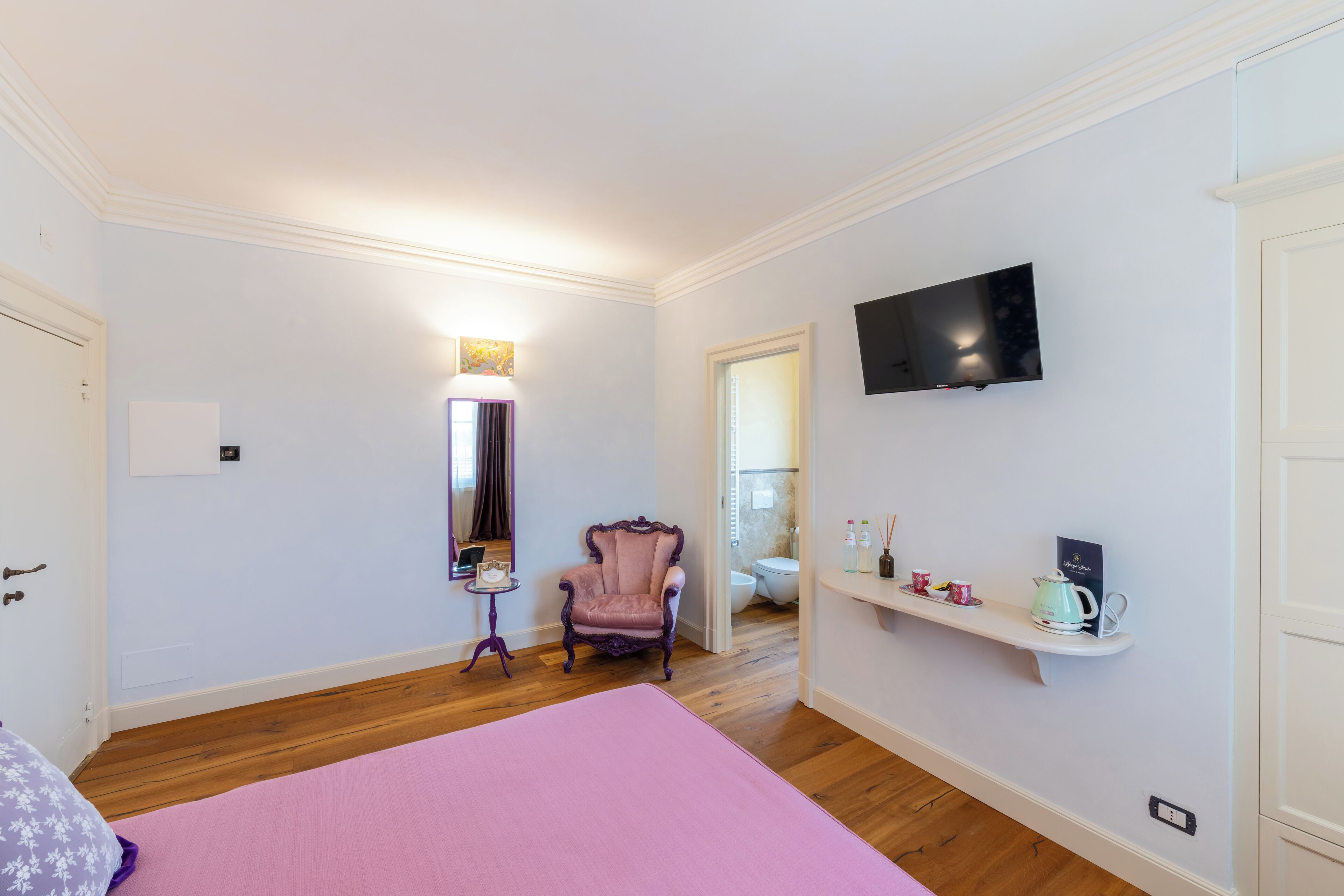 Un dormitorio con piso de madera, colcha rosa, sillón morado, una mesita de noche con lámpara, televisor en la pared y estante con artículos de tocador.