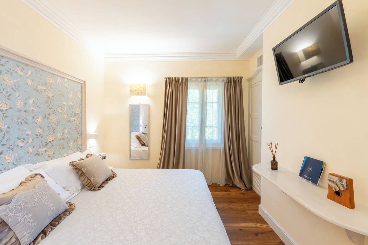 A bedroom with a large bed, a flat-screen TV mounted on the wall, a bedside table with a lamp, a mirror, and a window with curtains.