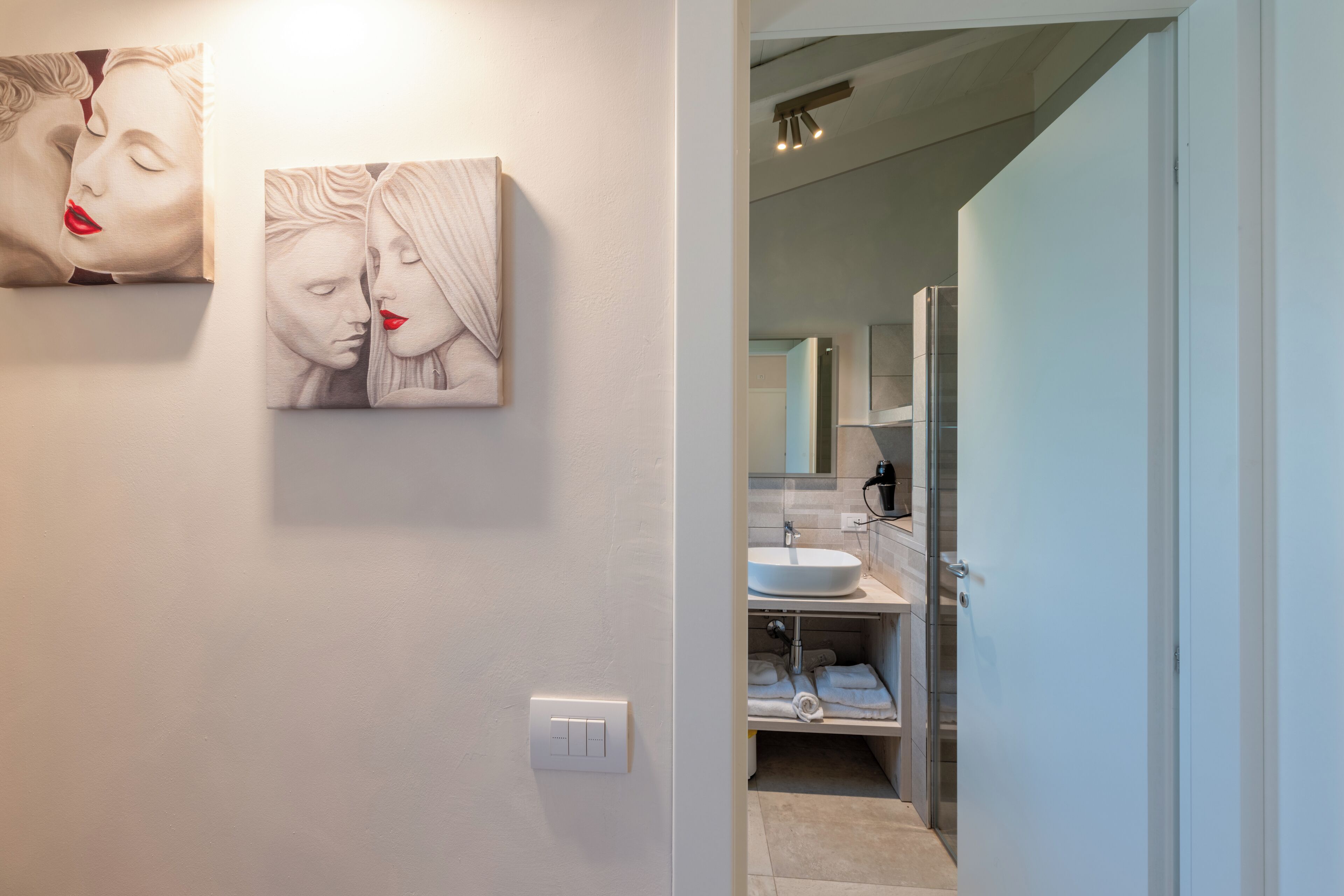 Une salle de bain moderne avec un lavabo blanc, un miroir et un porte-serviettes. Deux œuvres d’art murales représentant des visages abstraits y sont exposées.