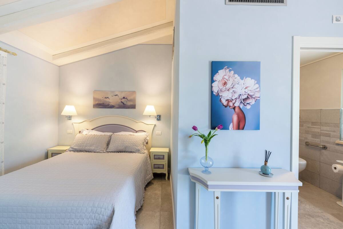 A bedroom with a bed, bedside lamps, a nightstand, a wall-mounted painting, and a small table with a vase of flowers.
