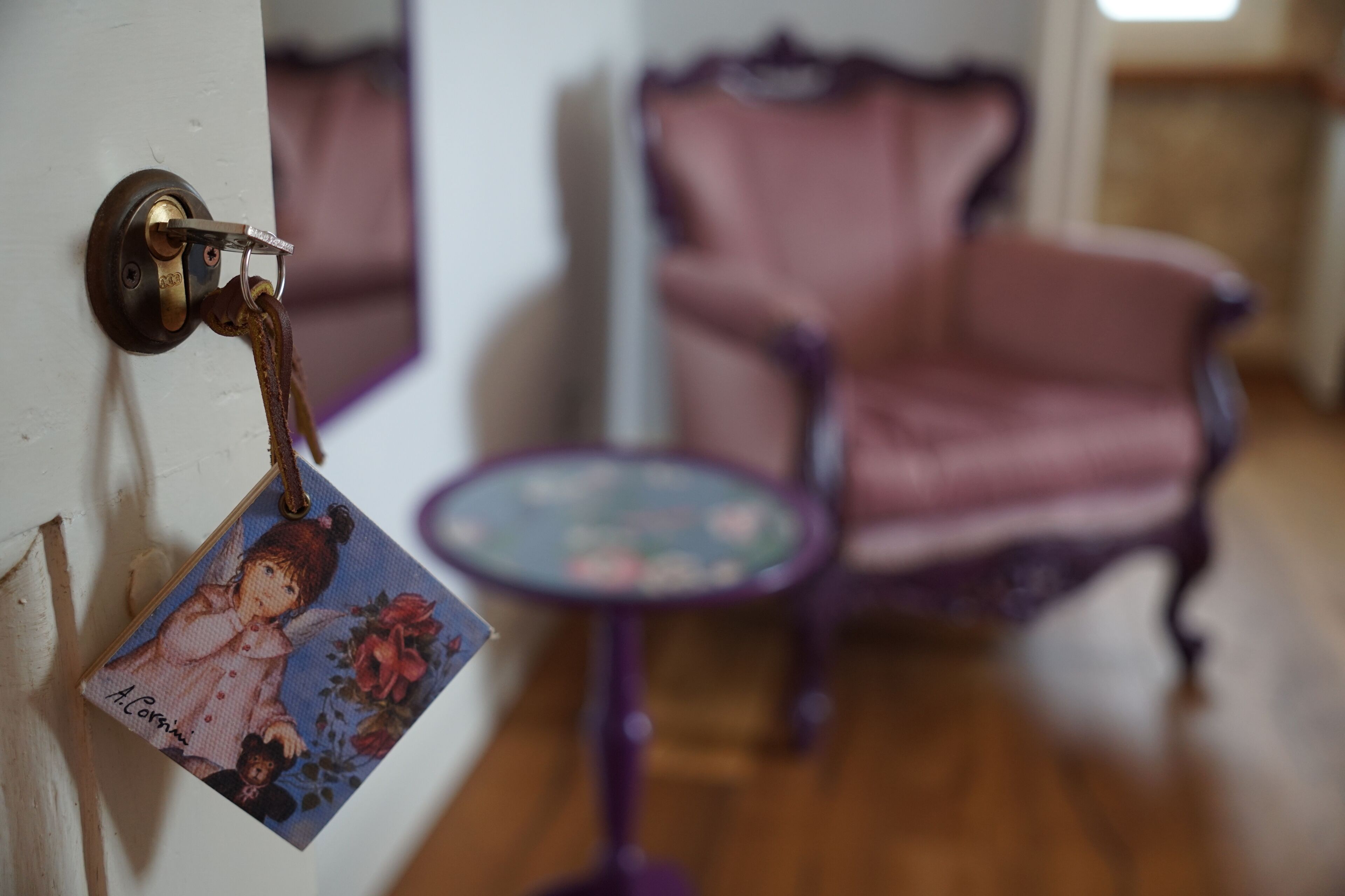 Une porte avec un trou de serrure et un porte-clés orné d’une photo. On aperçoit, à l’arrière-plan, une chaise rose et une petite table ronde à motifs floraux.