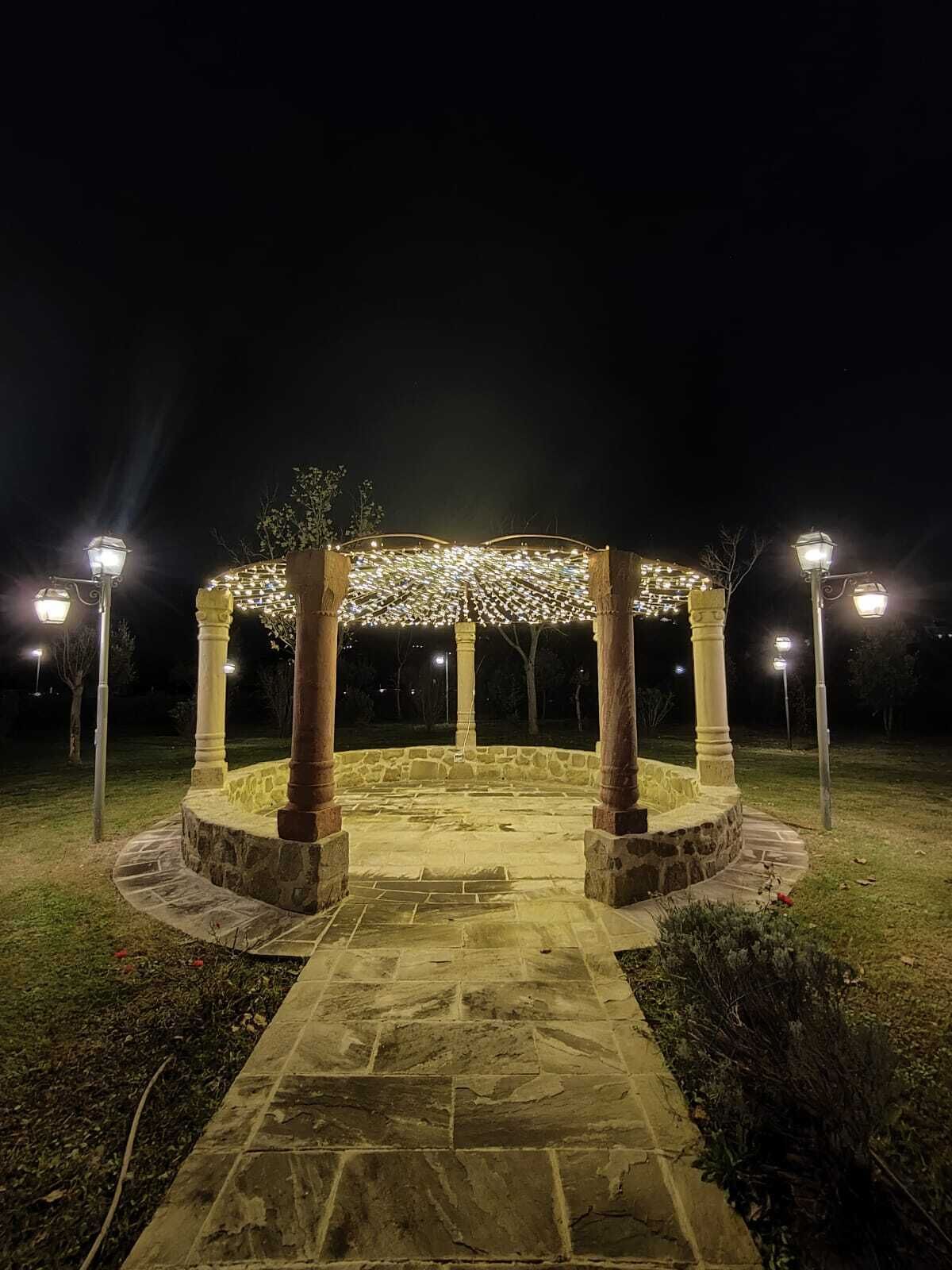 Un pabellón de piedra con techo iluminado, rodeado de un camino de piedra y faroles.