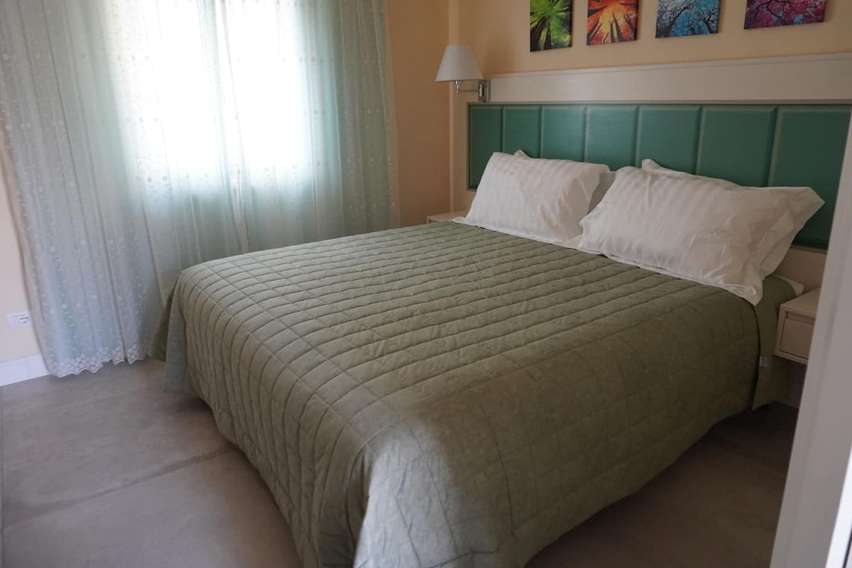 A neatly made bed with a green quilt, a white pillow, and a teal headboard in a room with a light-colored wall and a window with sheer curtains.