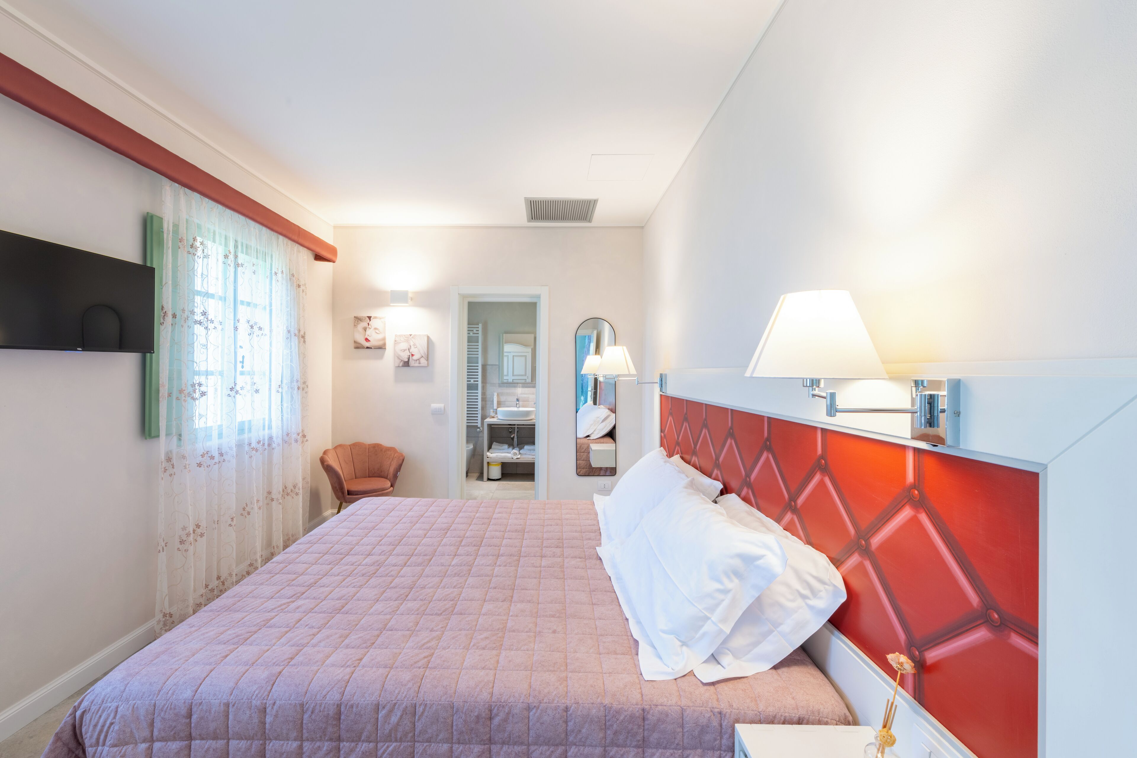 A hotel room with a bed, a chair, a nightstand, a mirror, and a bathroom visible through an open door.