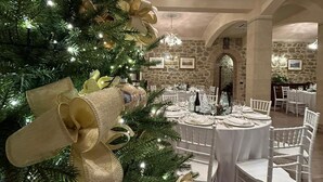 Un árbol de Navidad adornado con ornamentos dorados y lazos, una araña y un comedor con mesas redondas y sillas blancas.