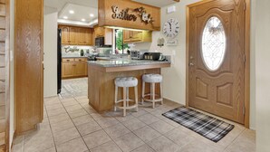 A kitchen with wooden cabinets, a central island with stools, a clock, and a door with a decorative glass panel. - Game Room + Bar + Lake Views | Gated Marina + Mini Golf | Naci Lakeview Retreat (Bradley)