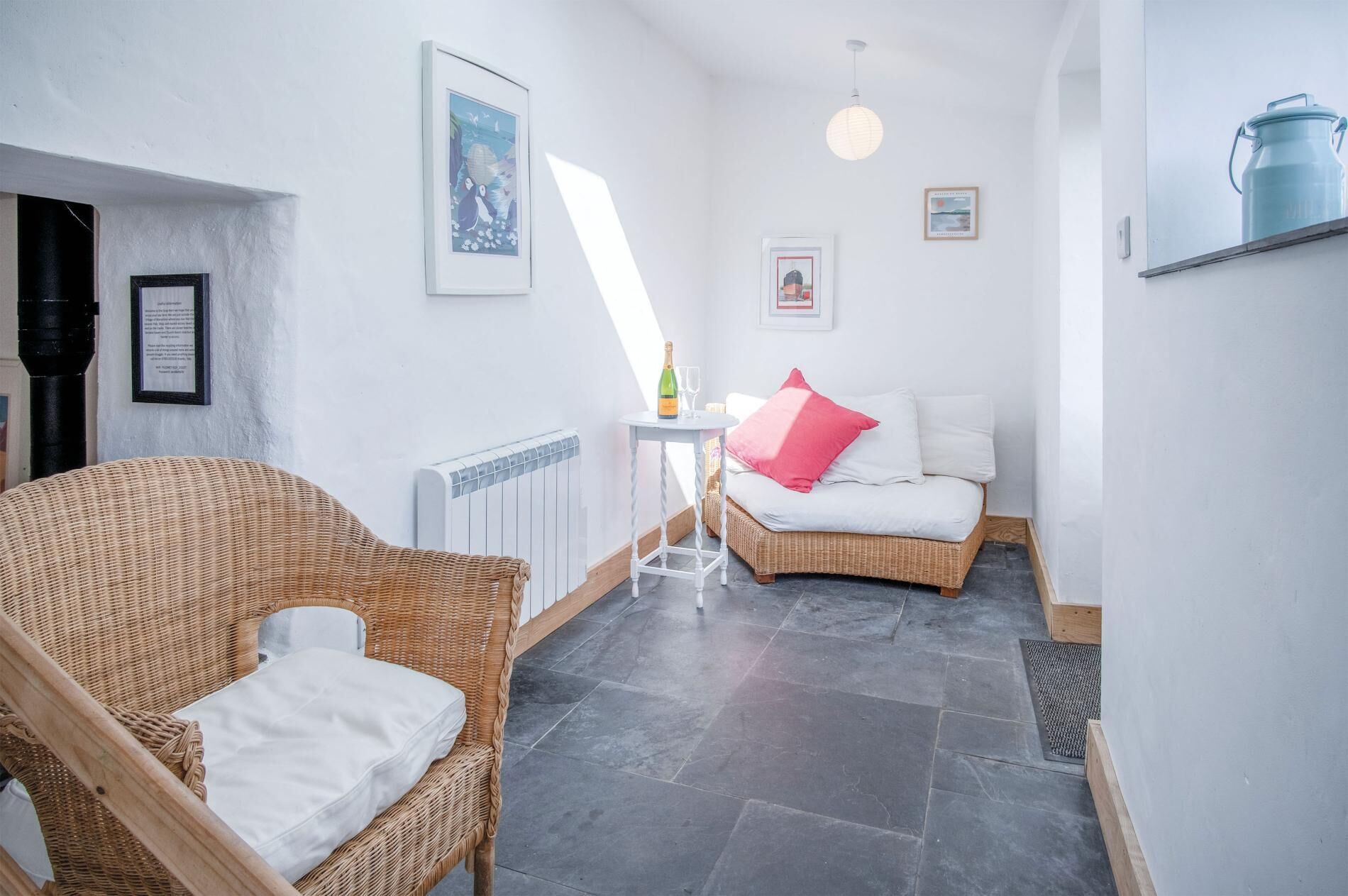 Une pièce chaleureuse avec un fauteuil en osier, un lit avec des oreillers blancs et roses, un radiateur et une petite table sur laquelle se trouvent une bouteille et des verres.