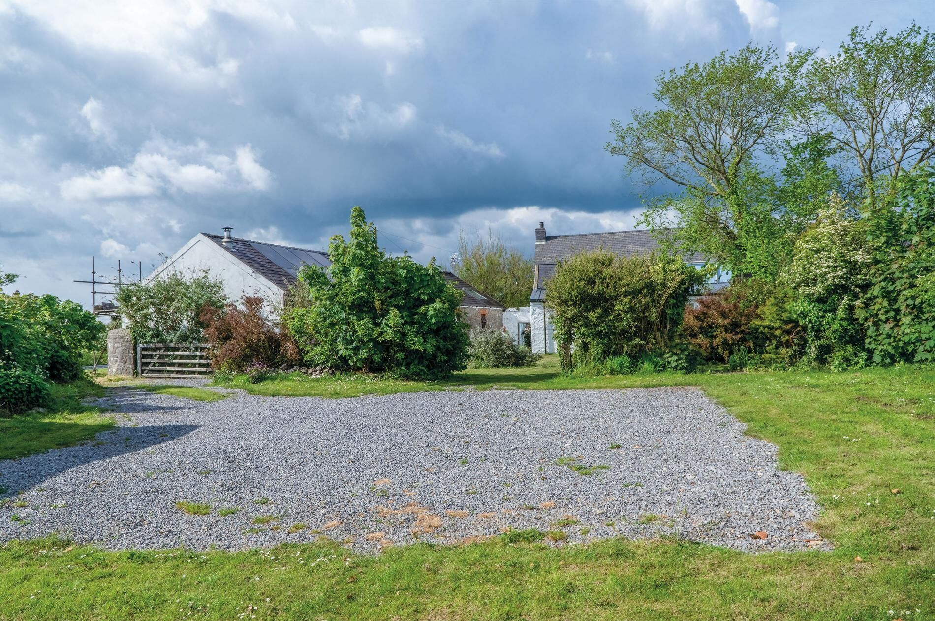 A gravel driveway leading to a white building with a sloped roof, surrounded by greenery and a cloudy sky.