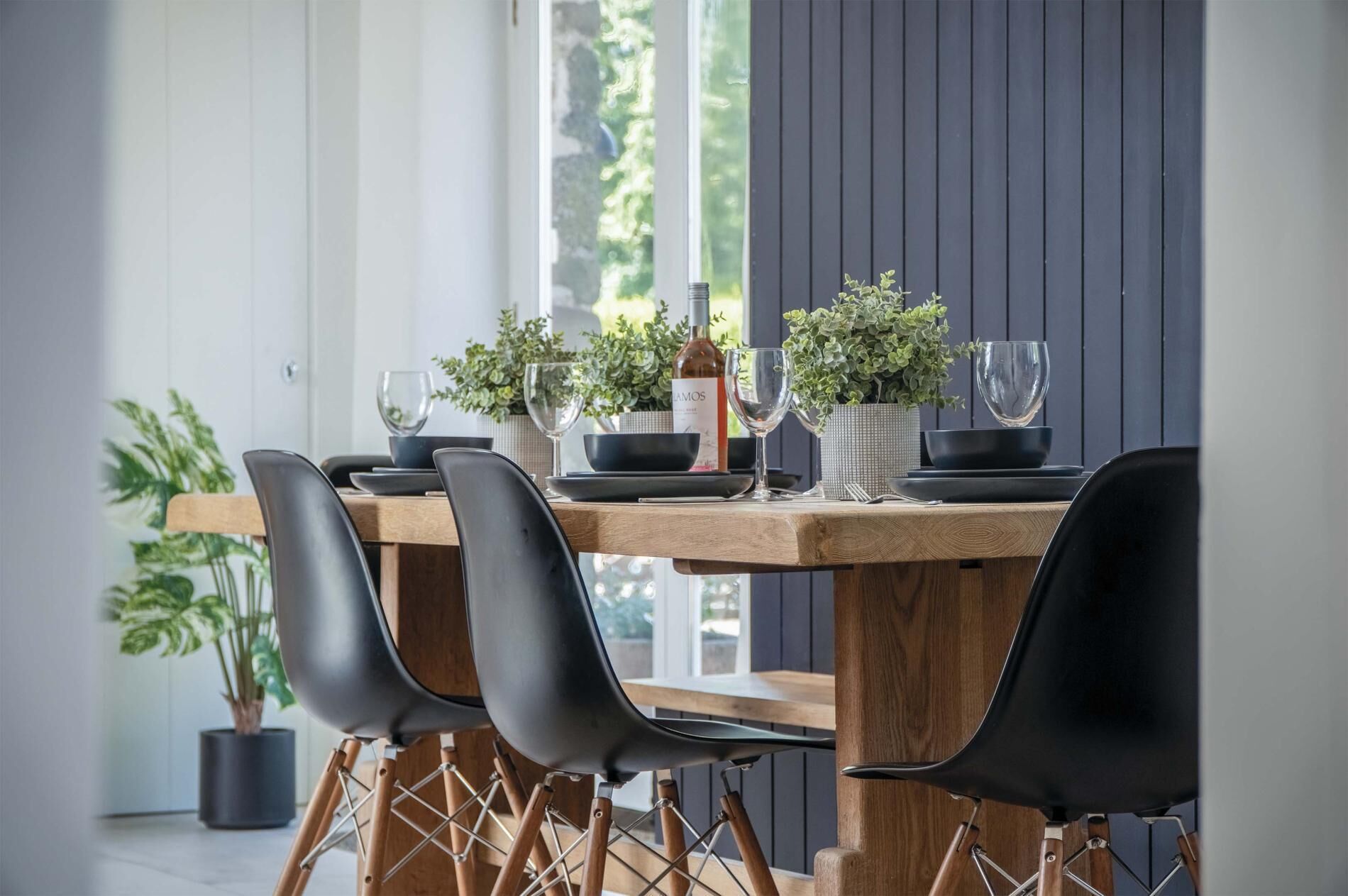 Een eetruimte met een houten tafel gedekt voor vier personen, omringd door zwarte stoelen met houten poten. Op de tafel staan potplanten en een fles wijn.