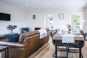 A modern living room with a brown leather sofa, a dining table set for four, and a flat-screen TV mounted on the wall. - Skomer Lodge - 2 Bedroom Lodge - Manorbier (Tenby)