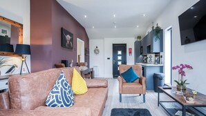 A modern living room with a brown leather sofa, a blue armchair, and a black rug. There is a dining area with a table and chairs, and a kitchen in the background.