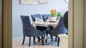 A dining area with a table set for four, surrounded by dark blue chairs. There is a painting on the wall above the table, and a potted plant with orange flowers on the table.