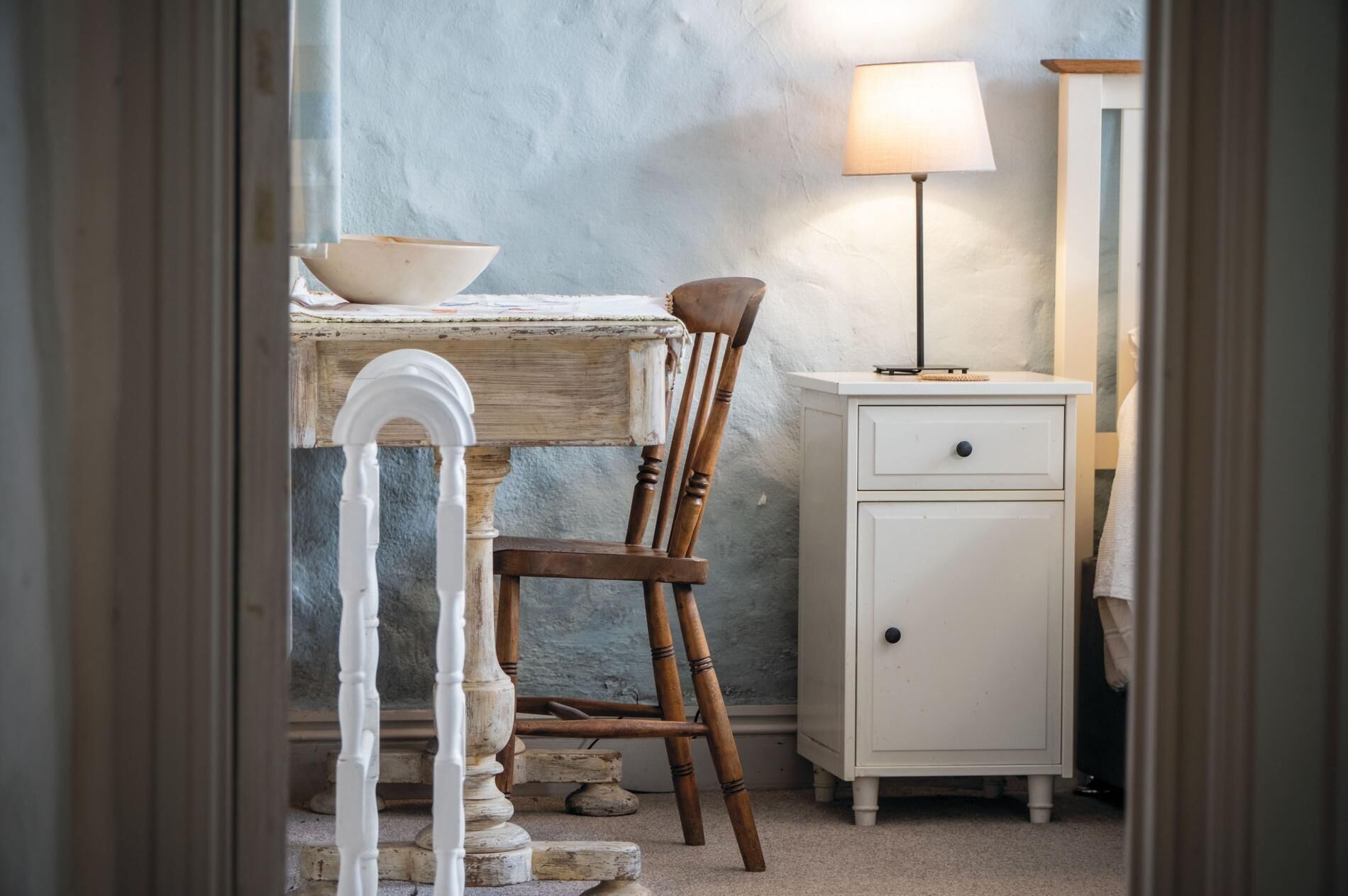 A rustic room with a wooden desk, a white chair, a small cabinet, and a lamp.