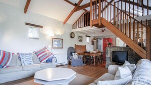 A spacious living room with a high ceiling, wooden beams, a staircase, a dining area, and a stone fireplace. - Haggard - 3 Bedroom Cottage - Manorbier (Tenby)