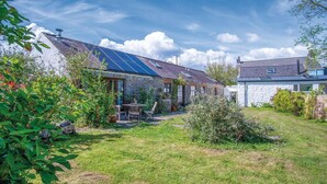 A stone house with a sloped roof, solar panels, and a garden with various plants. - Haggard - 3 Bedroom Cottage - Manorbier (Tenby)