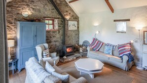 A cozy living room with a stone wall, a fireplace, a sofa, and a blue armchair. - Haggard - 3 Bedroom Cottage - Manorbier (Tenby)