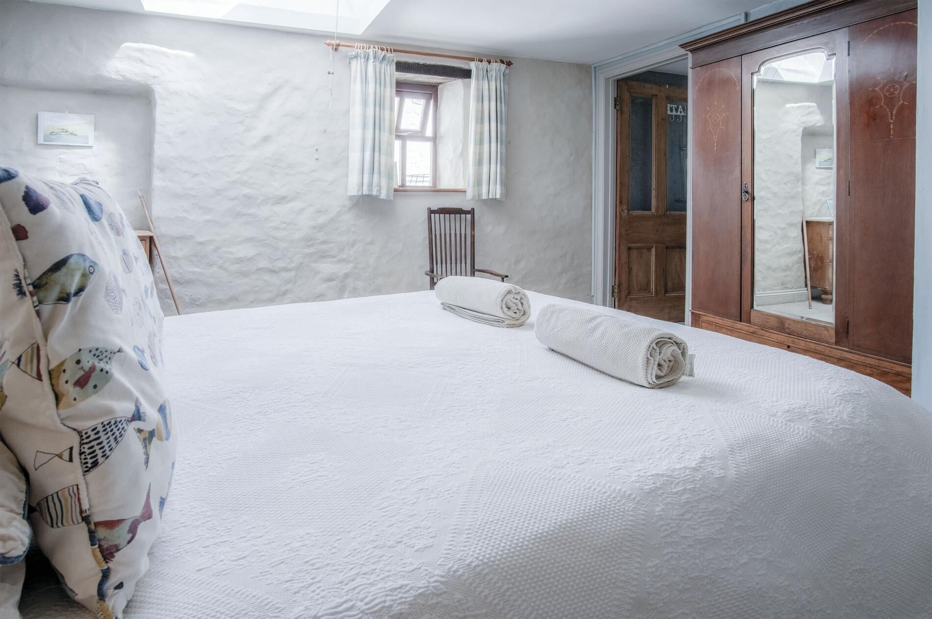 A bedroom with a large bed, a wooden chair, a window with curtains, and a wardrobe.