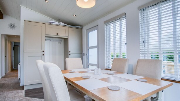Une cuisine moderne avec des meubles blancs, une table à manger en bois et quatre chaises, ainsi que de grandes fenêtres équipées de stores.