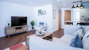 A modern living room with a flat-screen TV, a white sofa, a coffee table, and a dining area with pendant lights. - 25 Captains Walk - 1 Bedroom Apartment - Saundersfoot (Saundersfoot)