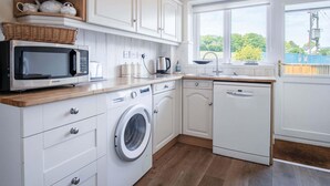 A kitchen with white cabinetry, a washing machine, a microwave, and a window with a view of greenery. - Clover Cottage - 2 Bedroom Cottage - Begelly (Kilgetty)