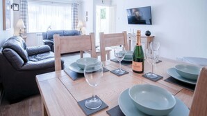 A dining area with a wooden table set for two, including wine glasses, plates, and a bottle of wine. There is a leather sofa and a television in the background. - Clover Cottage - 2 Bedroom Cottage - Begelly (Kilgetty)