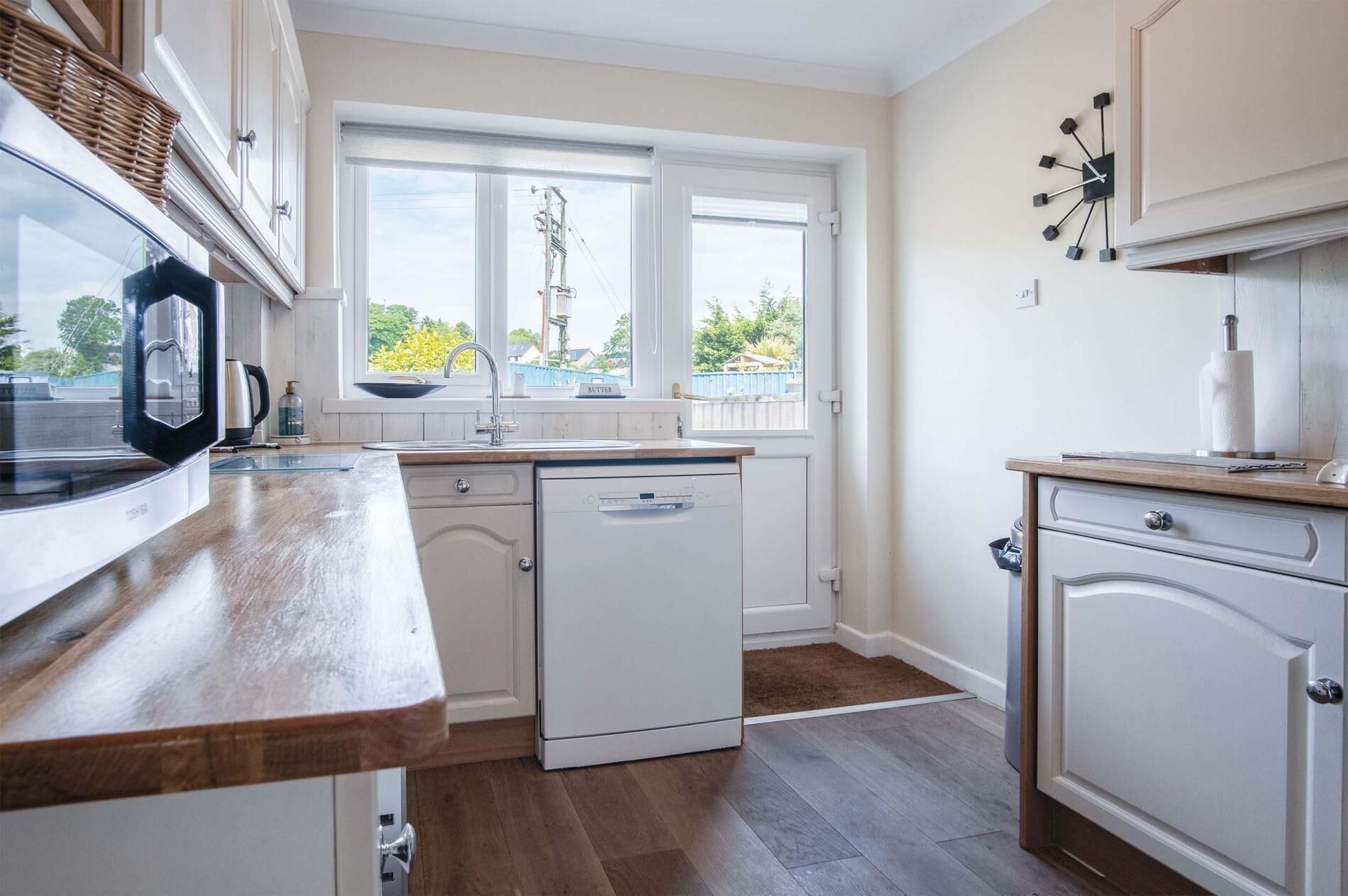 A kitchen with a wooden countertop, white cabinets, a built-in dishwasher, and a window with a view of greenery.