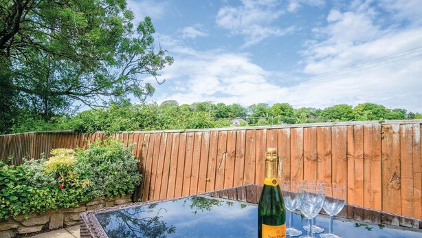 A bottle of champagne and glasses on a glass table outdoors with a wooden fence and greenery in the background. - Clover Cottage - 2 Bedroom Cottage - Begelly (Kilgetty)