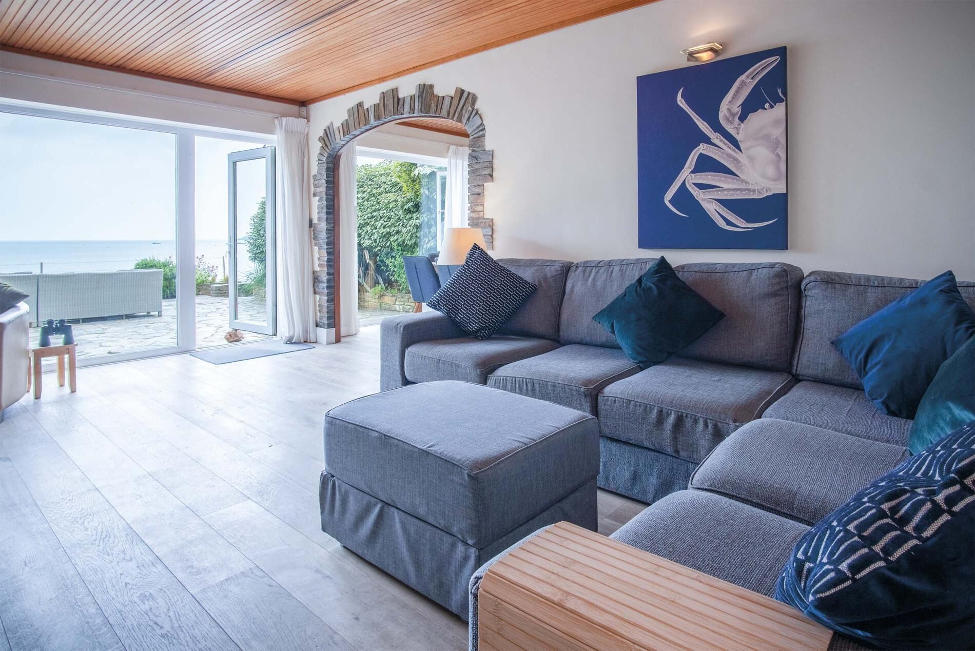 Un salon moderne avec un canapé gris, un pouf et une table basse en bois. Une grande fenêtre offre une vue sur la mer.