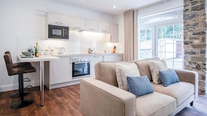 A modern kitchen with white cabinetry, a built-in oven, and a microwave. There is a dining area with a table and chairs, and a large window with a stone wall. - The Carthouse - 1 Bedroom Cottage - Princes Gate (Narberth)