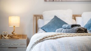 A neatly made bed with a white and blue pillow arrangement, a folded towel, and a bedside lamp on a wooden nightstand. - The Carthouse - 1 Bedroom Cottage - Princes Gate (Narberth)