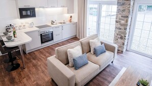 A modern kitchen with white cabinets, a built-in microwave, and a dining area with a table and chairs. - The Carthouse - 1 Bedroom Cottage - Princes Gate (Narberth)